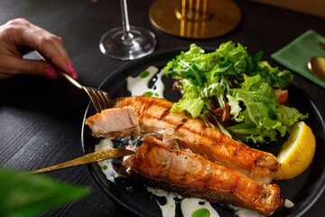 One piece of baked salmon grilled pepper lemon and salt on a black plate with lettuce leaves on a stone background