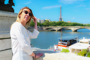 Naklejka premium Portrait of a pretty blonde woman in town by the water, standing on a beautiful historic bridge. Eiffel tower and Seine river in the background. 