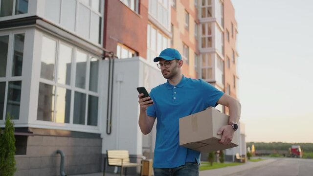 A male mail carrier with round glasses carries boxes for delivery to customers and looks at his mobile phone. Search through the mobile phone for the delivery address. Payment terminal.
