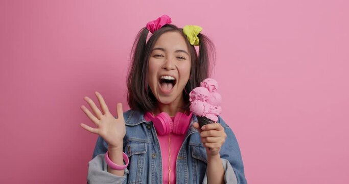 Emotional Asian girl screams loudly keeps palm raised taste delicious ice cream dressed in fashionable denim jacket being very glad to meet old friend poses with headphones against pink wall