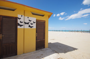 In the foreground the changing room with yellow wall and black butterflies on a white background, in the distance the beach before summer and the sea.