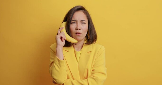 Irritated Asian woman pretends having telephone coversation holds banana near as if phone screams angrily has quarrel with someone looks with grumpy expression isolated over yellow studio wall