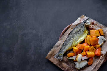 Trout baked whole in spices with vegetables-pumpkin and onion. In baking paper on a wooden board. Black background, copy space