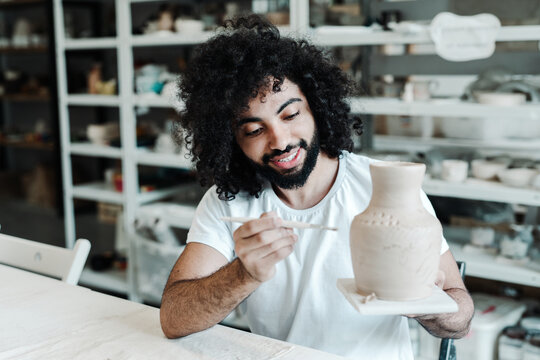 Happy african american man in a t-shirt holds a clay vase in his hand and paints it with a brush