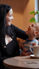 Close up portrait of a little boy with special needs and mom laughing at a table in a cafe, lifestyle. Mom's love for her child, inclusion.Happy disability kid concept