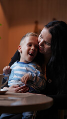 Close up portrait of a little boy with special needs and mom laughing at a table in a cafe,...
