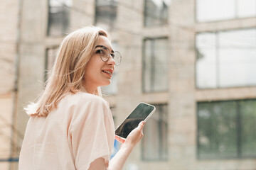 Back View of Elegant Woman in Eyeglasses Holding Smartphone in Hand and Smiling, Stylish Blonde Girl with Glasses Standing Outdoors on the Street Looking Away