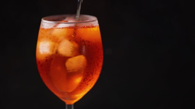 Cocktail aperol spritz on black background. Spoon slowly mixes beverage. Glass of summer orange cocktail. Slow motion. Copy space
