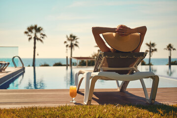 Rear view woman wear hat sunbathing on deckchair on poolside - obrazy, fototapety, plakaty