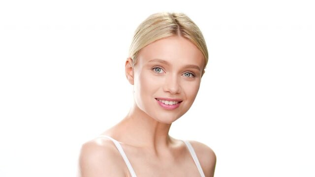 Slim young beautiful Caucasian woman with light hair in white top turns to the left touching jaw line and smiling wide for the camera on white background | Moisturizer commercial