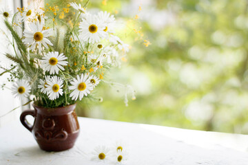 a bouquet of daisies, wildflowers and greens in a clay vase in the form of a mug on a wooden white windowsill against the window. Postcard.
