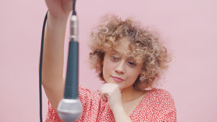 Attractive young girl holding a microphone through its wire. The mic is hanging upside down. Girl thinking. Dressed in a pink casual top. Isolated over pink background studio.