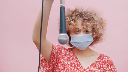 Girl wearing a mask and holding a microphone upside down through its wire. Serious looking girl with curly hair and dressed in a pink casual top. Isolated over pink background studio. 