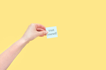 The hand holds a sticky note with inscription Trust yourself