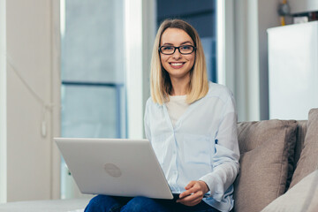 Portrait of a blonde woman looking at the camera sitting at home on the couch and holding a laptop