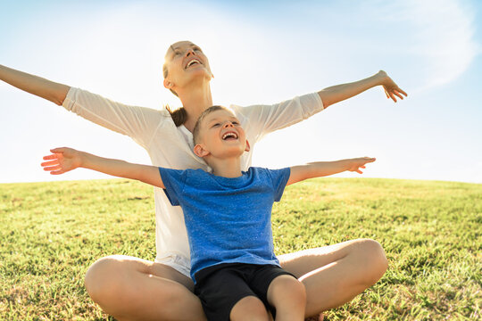 Portrait of mother and child sitting in a grass field with arms out to the sky feeling happy and free. 