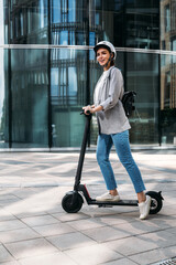 Young happy woman in casuals driving electric push scooter. Businesswoman in cycling helmet standing on e-scooter.