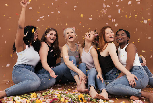 Group of happy women sitting on the brown background while petals from flowers falling. Laughing females of different races and ages under petals from flowers.