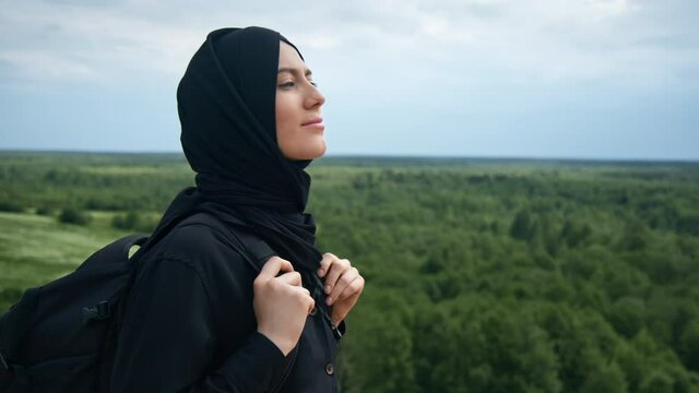 Smiling Muslim backpacker female admiring nature landscape from top of mountain travel vacation