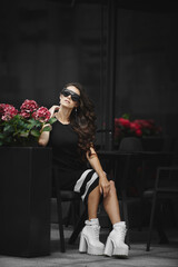 Fototapeta na wymiar A fashionable young woman in trendy sunglasses and stylish dress sitting at the cafe table outdoors