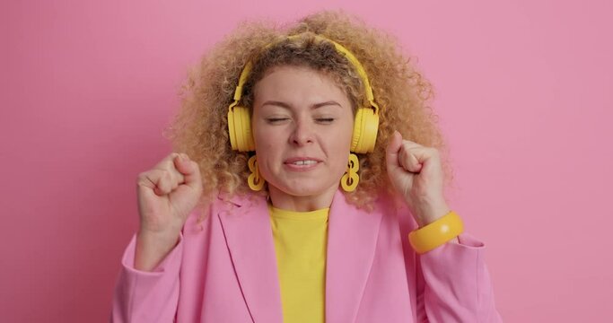 Cheerful curly haired woman puts all efforts in making wish crosses fingers has faith for better clenches fists with triumph smiles broadly wears wireless headphones on ears dressed formally