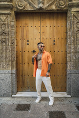 Hot caucasian man with arm tattoos wearing an orange shirt and white pants posing at the entrance