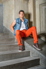 A young guy, sitting outside and against a wall, is looking at you.