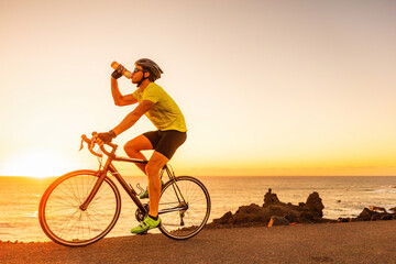 Cycling sports athlete cyclist man drinking water bottle while biking on road bike at sunset. - obrazy, fototapety, plakaty
