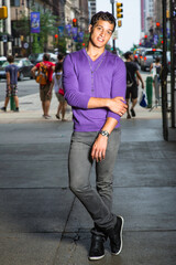 Wearing a purple sweater, a necklace,  black pants and black shoes, a young handsome guy is fashionably standing in a gateway to a busy street..