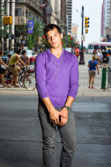 Wearing a purple sweater, a necklace,  black pants and black shoes, a young handsome guy is  tired and standing in a gateway to a busy street..