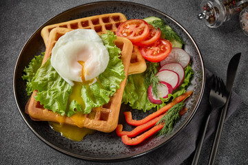 Tasty waffles with poached egg and vegetables on black plate on grey background. Top view.  Table setting