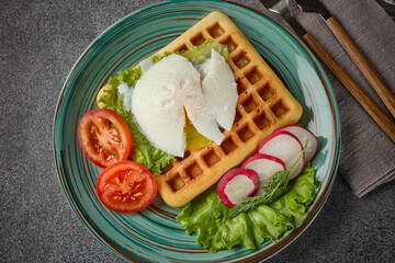 Tasty waffles with poached egg and vegetables on blue plate on grey background. Top view. Table setting