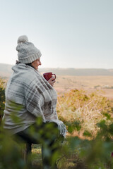 Latin woman sitting on a rustic chair, wearing a warm hat and scarf, drinking coffee while contemplating the landscape and smiling on a cold morning or afternoon. Vertical shot.