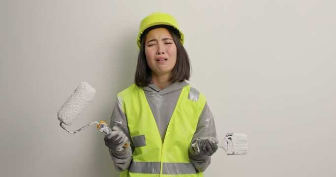 Frustrated Asian female construction worker feels upset as doesnt want to work holds painting roller brush wears protective helmet and uniform isolated over white background busy with refurbishment