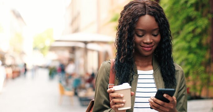 Portrait of the attractive young multiracial woman talking smartphone and drinking coffee outdoors. Beautiful girl using mobile phone while making call at the evening street with blurred background