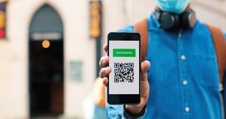Portrait view of man wearing protective mask calmly looking at camera and showing vaccine passport at his smartphone while standing at empty street