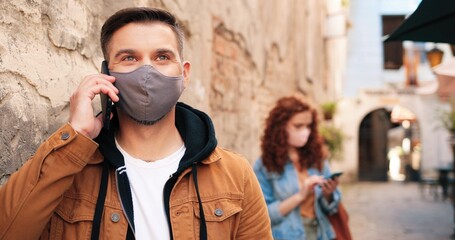 Waist up portrait view of the brunette caucasian man wearing protective face mask having conversation via smartphone on urban space. Outbreak of coronavirus COVID-19, pandemic and health care concept