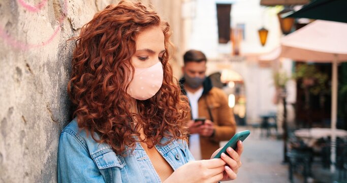 Waist up portrait view of the ginger caucasian girl wearing protective face mask using her smartphone on urban space at sunset. Outbreak of coronavirus COVID-19, pandemic and health care concept