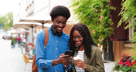 Stylish multiracial man and his girlfriend using his smartphone while walking down the street. Handsome man chatting with his friend during a city walk in summer
