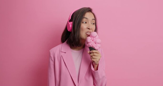 Displeased brunette young Asian woman licks ice cream which doesnt taste good smirks face wears wireless headphones fashionable jacket isolated over pink background. I dont like this flavor.