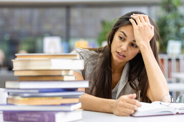 Beautiful caucasian female student resentful of the large amount of educational material sitting in the library