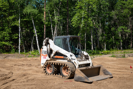CROW WING CO, MN - 6 JUN 2021: A Bobcat S185, white compact front end loader, with skid steer tracks, sits on dirt at a tree lined empty building lot, in preparation for new home construction.