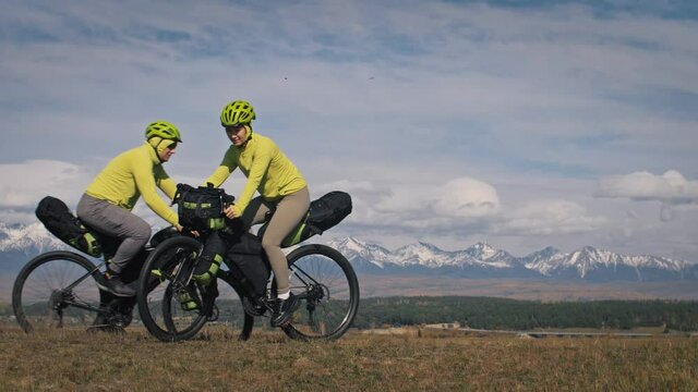 The man and woman travel on mixed terrain cycle touring with bikepacking. The two people journey with bicycle bags. Sport bikepacking, bike, sportswear in green black colors. Mountain snow capped.