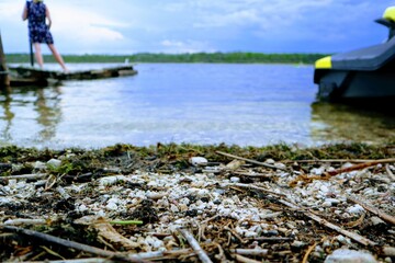 Fototapeta premium Close-up of a lake shore covered with pebbles, grass and algae. A girl standing on the pier and a jet ski in the background.