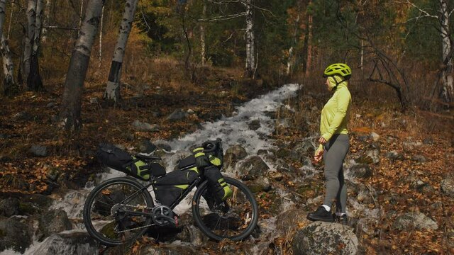 The woman travel on mixed terrain cycle touring with bikepacking. The traveler journey with bicycle bags. Sportswear in green black colors. The trip in magical autumn forest, river stream.