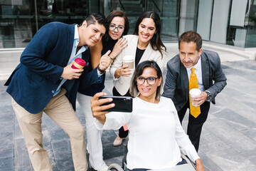 Hispanic transgender businesswoman in wheelchair taking selfie with co-workers on coffee break, in disability concept and disabled people
