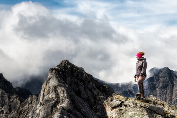 Young woman with a backpack stands on the edge of a cliff and looks at the sky with clouds in the mountains