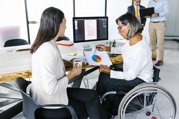 Hispanic transgender businesswoman in wheelchair and co-worker holding cup of coffee reviewing...