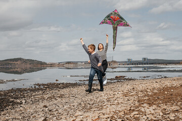 Mom and son run along the river bank with a kite.