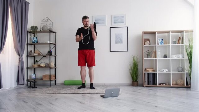 Fitness beginner. Chubby man. Healthy sport. Online training. Lockdown lifestyle. Puzzled middle-aged guy looking exercises on laptop studying resistance band light room interior.